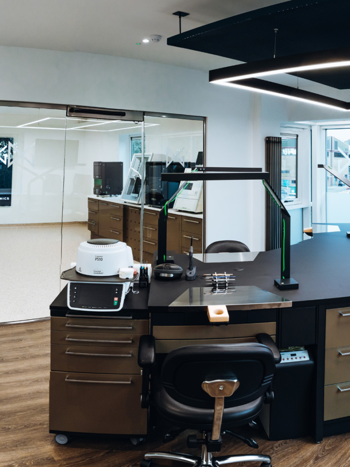A large office room inside of a dental laboratory with multiple work and computer stations on a large dark work top in the centre of the room. Behind a glass window there are several high-tech dental laboratory machines.