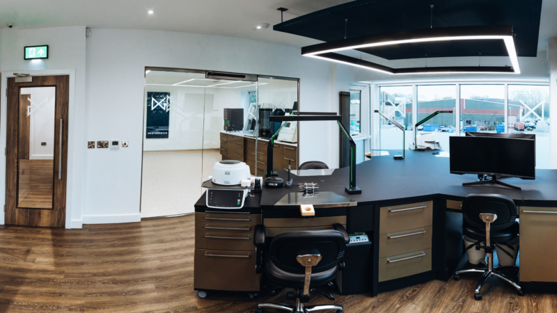 A large office room inside of a dental laboratory with multiple work and computer stations on a large dark work top in the centre of the room. Behind a glass window there are several high-tech dental laboratory machines.