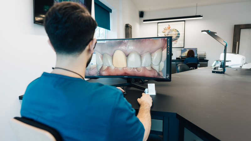 The back of a lab technician wearing blue scrubs looking at a 3D render of a seth of teeth on his computer screen at a dark work station.