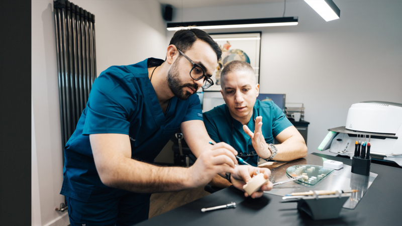 Two male dental lab technicians in blue scrubs looking over and discussing a piece of prosphetic work they're working on.