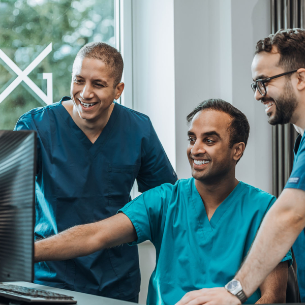 Three lab technicians in blue scrubs smiling and looking at a computer screen together.
