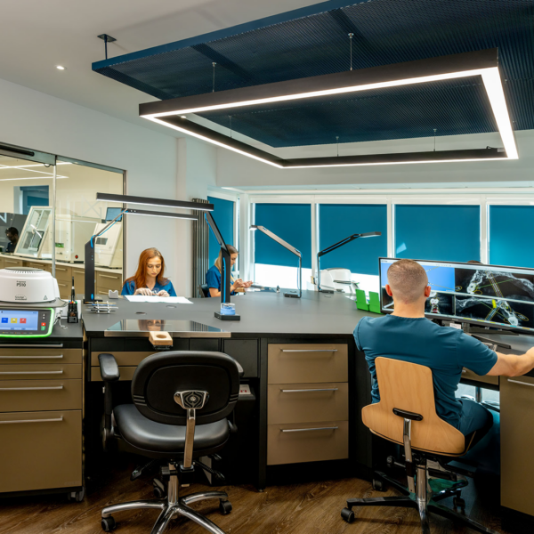 Three lab technicians sat at a large work table with multiple work stations. Some are working on prosphetics and one lab technician is looking at x-rays on a monitor. There are machines in the background behind a glass door.