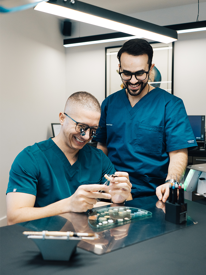Two lab technicians in blue scrubs working on a tooth prosphesis. One of the technicians is wearing magnifying glasses while the other stands next to him looking at the prosphetic.