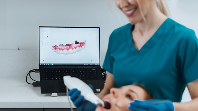 A dentist in blue scrubs is placing a machine inside a patients mouth while a digital scan of the top set of their teeth shows up on the laptop screen behind them.